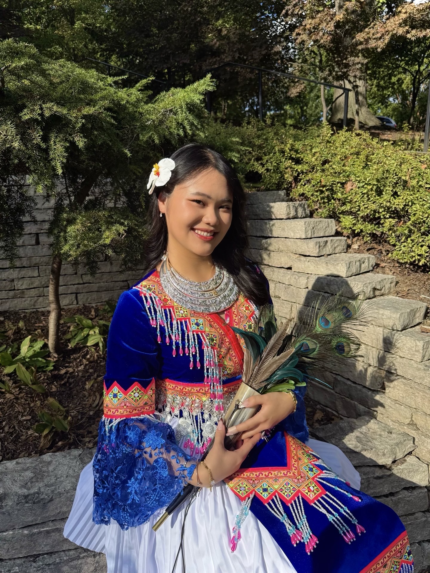 Cindy Vue, a young woman of Hmong descent, is smiling at the camera. She is wearing traditional Hmong attire, which is intricately beaded with bold colors. 
